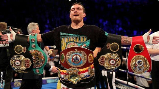 Oleksandr Usyk posing triumphantly while holding all four major world heavyweight championship belts, including the WBC, WBA, IBF, and WBO titles.