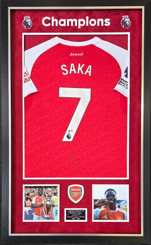 Front view of a luxury framed 2025/26 Arsenal home shirt signed on the back by Bukayo Saka, featuring CHAMPIONS top header, celebration images, and an engraved plaque.