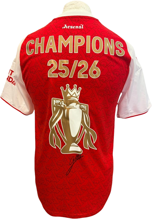 The reverse of the red and white Arsenal 2025/26 home jersey showcasing Gabriel Martinelli's hand-signed autograph on the fabric next to the gold Premier League trophy and Champions 25/26 printing.